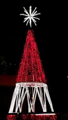 Estructura navideña en forma de árbol iluminada con luces rojas y blancas sobre fondo negro. La composición destaca por su contraste y diseño moderno, ideal para representar iluminación artística y decoración navideña contemporánea.