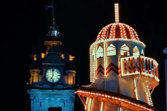 Vista nocturna de Edimburgo en Navidad. A la izquierda se alza el Balmoral Hotel con su torre del reloj iluminada en tonos azules y dorados, marcando la hora sobre la fachada de piedra. A la derecha, una gran atracción de feria navideña brilla con luces cálidas rojas, blancas y amarillas, destacando su estructura decorada y su cúpula luminosa. El cielo oscuro enmarca el contraste entre la arquitectura histórica de la ciudad y el ambiente festivo de las celebraciones navideñas, evocando el espíritu invernal y acogedor de Edimburgo durante las fiestas.