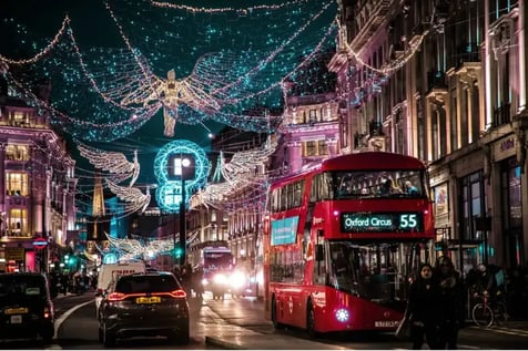Vista nocturna de Oxford Street en Londres durante la Navidad, con un autobús rojo de dos pisos circulando bajo una espectacular decoración de luces en forma de ángeles y alas luminosas suspendidas sobre la calle, rodeado de edificios históricos iluminados y tráfico urbano.