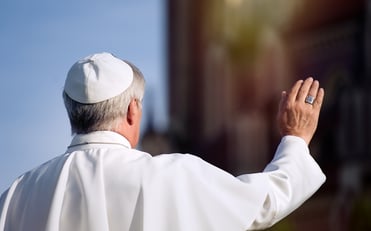 Figura del papa vista de espaldas, con sotana blanca, levantando la mano en gesto de saludo o bendición.