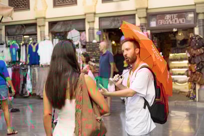 Guía turístico con paraguas naranja de My Top Tour hablando con una visitante en una calle comercial concurrida.