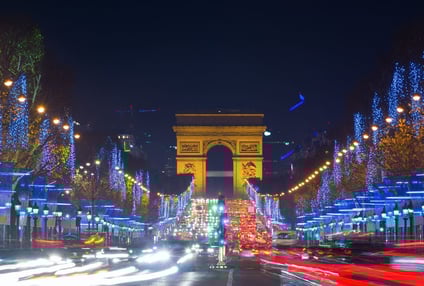 Vista nocturna del Arco del Triunfo en París durante la Navidad, con los Campos Elíseos iluminados por luces azules y doradas en los árboles, tráfico urbano en movimiento creando estelas de luz y una atmósfera festiva que combina el dinamismo de la ciudad con la elegancia parisina en época navideña.  Select 50 more words to run Humanizer.