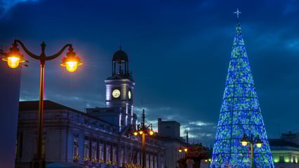 Gran árbol de Navidad iluminado con luces azules y verdes en una plaza urbana al anochecer. Al fondo se distinguen edificios históricos y farolas encendidas, creando una escena navideña elegante y representativa de la decoración monumental de las ciudades.