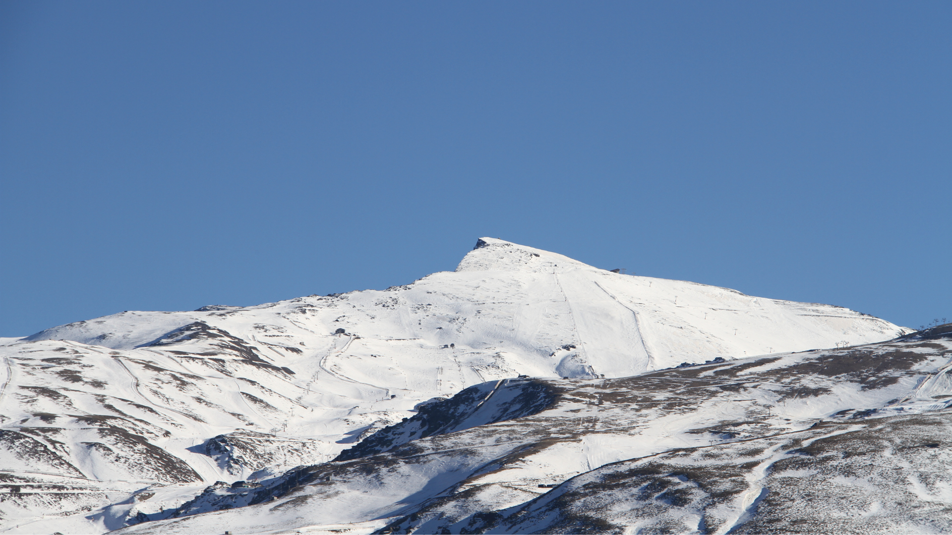 Vista panorámica de una gran montaña cubierta de nieve bajo un cielo completamente despejado y azul intenso. La imagen muestra las laderas blancas con algunas zonas rocosas visibles, transmitiendo sensación de invierno, frío y naturaleza en estado puro. Ideal para representar paisajes de alta montaña o destinos de nieve.