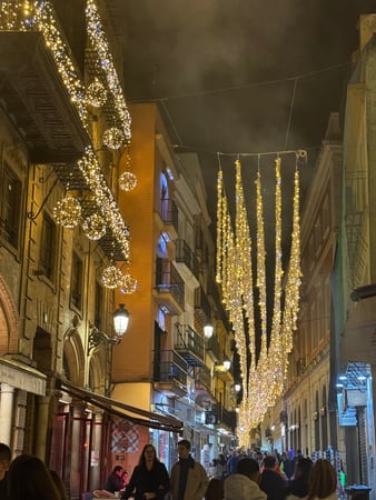 Calle céntrica de una ciudad al anochecer, decorada con abundante iluminación navideña en tonos cálidos. Guirnaldas y adornos luminosos cuelgan entre los edificios mientras peatones pasean, creando un ambiente festivo, acogedor y muy animado propio de la Navidad urbana.