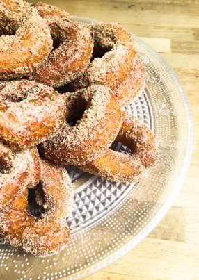 Primer plano de varias rosquillas caseras cubiertas de azúcar, colocadas sobre una bandeja de cristal transparente. Se aprecia la textura crujiente del exterior y el aspecto artesanal del dulce, evocando tradición gastronómica, repostería casera y sabores típicos de invierno.