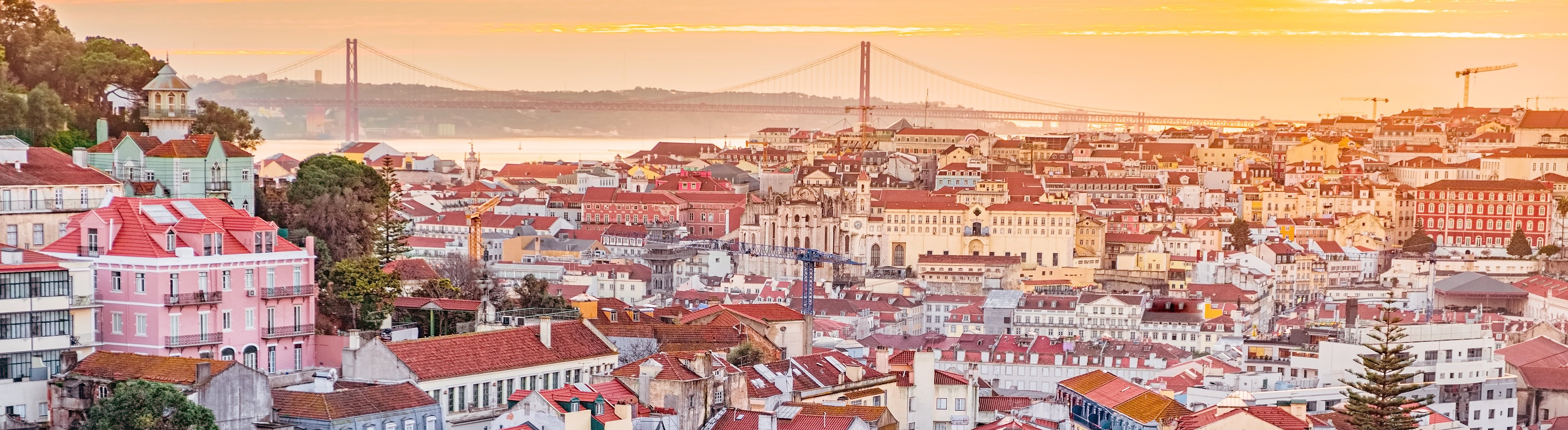 Vista panorámica del barrio de Alfama y el centro de Lisboa al atardecer, con el puente 25 de Abril al fondo sobre el río Tajo.