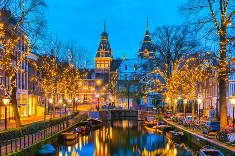 Canales de Ámsterdam en Navidad al atardecer, con árboles sin hojas cubiertos de luces cálidas reflejadas en el agua, bicicletas aparcadas junto a las orillas y edificios históricos iluminados que crean un ambiente acogedor y festivo.