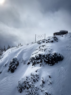 Montaña nevada con los anillos olímpicos instalados en la cima, bajo un cielo nublado y un entorno alpino invernal.