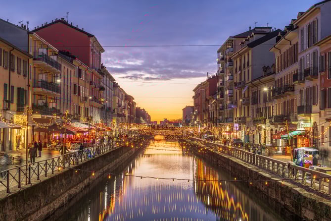 Canal de Navigli en Milán al atardecer, con edificios de colores, luces decorativas reflejadas en el agua y ambiente urbano animado.