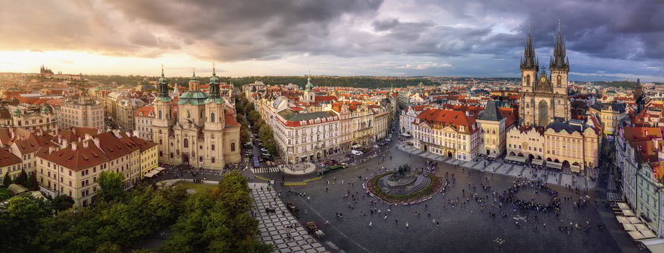 Panorama de la Plaza de la Ciudad Vieja de Praga con edificios históricos, cúpulas verdes y torres góticas bajo un cielo dramático.