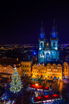 Vista nocturna de la Plaza de la Ciudad Vieja de Praga en Navidad, con un gran árbol decorado con luces, mercados navideños llenos de gente y la Iglesia de Nuestra Señora de Týn iluminada dominando el paisaje urbano.