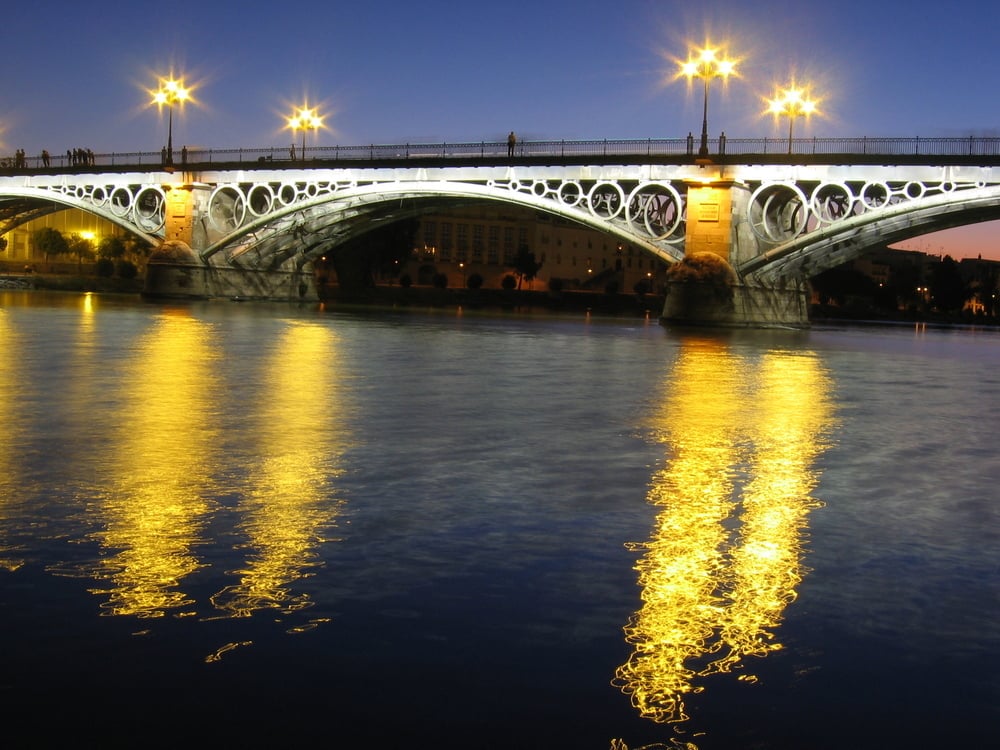 Imagen del puente de Triana de Sevilla de noche cuya iluminación se refleja en el Río Guadalquivir