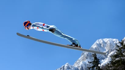 Saltador de esquí en pleno vuelo durante una prueba de salto, con traje aerodinámico y montañas nevadas al fondo.