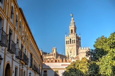 La Giralda y la Catedral de Sevilla al fondo, vistas desde una calle con fachadas amarillas y balcones de hierro, bajo cielo azul.