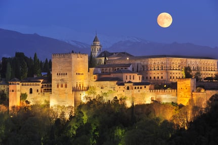 La Alhambra de Granada iluminada de noche, con luna llena sobre Sierra Nevada al fondo.