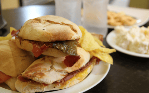 Bocadillo de jamón con pimiento y patatas chips en un plato, sobre una mesa de bar.