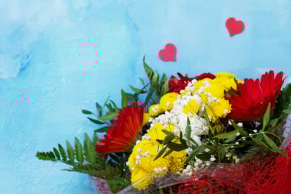 Ramo colorido de flores (gerberas rojas, crisantemos amarillos y paniculata blanca) envuelto en malla roja sobre fondo azul, con dos corazones decorativos en la parte superior.